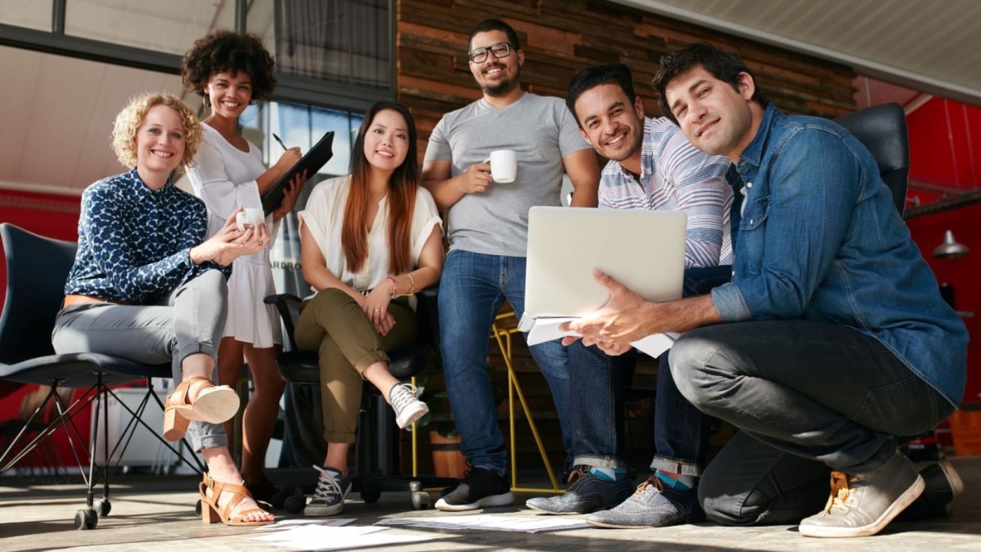 A diverse, friendly group of colleagues smiles together in a casual office setting. They sit and crouch closely, holding a laptop, coffee mugs, and notebooks, showing a warm, collaborative team atmosphere.