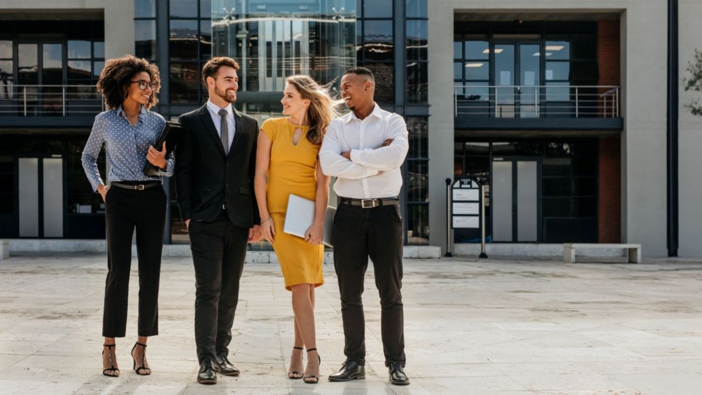 A group of four diverse, professionally dressed colleagues walks together outside a modern office building. They are smiling and engaged in conversation, projecting a sense of teamwork, confidence, and collaboration. The individuals are dressed in business attire, with one person holding a notebook, emphasizing a professional and motivated atmosphere.