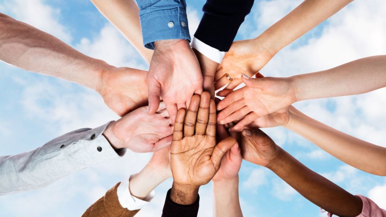 The image shows a group of diverse hands joined together in a circle against a blue sky background, symbolizing unity, teamwork, and diversity. The hands vary in skin tone, suggesting inclusivity and collaboration among people from different backgrounds.