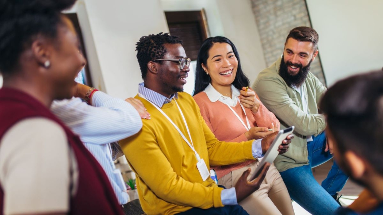 A diverse group of colleagues sitting together in a casual office setting, engaged in a lively discussion and smiling, with one person holding a tablet.