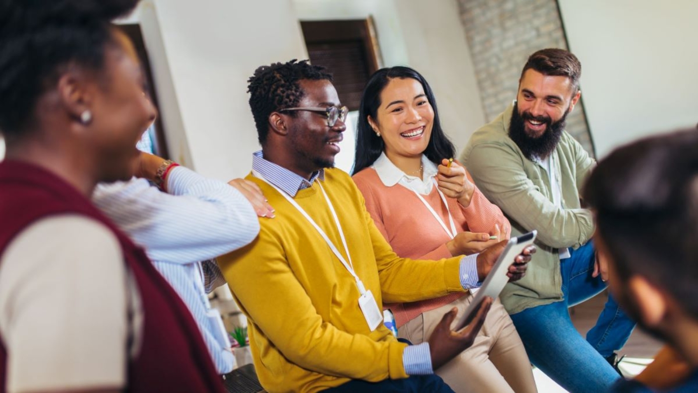 A diverse group of colleagues sitting together in a casual office setting, engaged in a lively discussion and smiling, with one person holding a tablet.