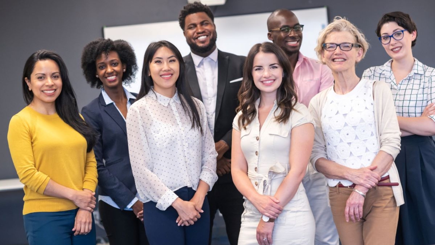 A diverse group of professionals standing together and smiling in a conference or office setting, representing teamwork and inclusivity.