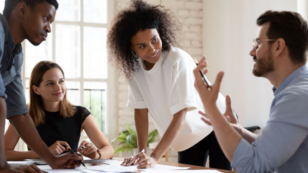 Office environment with four coworkers having a focused discussion, led by a woman standing at the table.