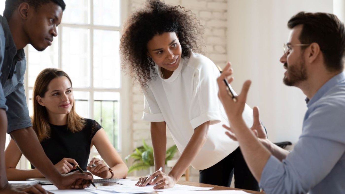 Office environment with four coworkers having a focused discussion, led by a woman standing at the table.