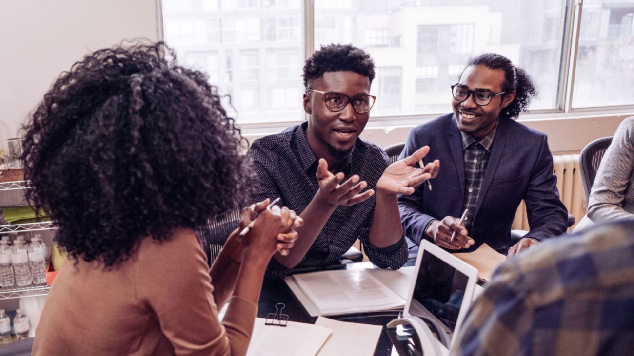 Diverse team collaborating around a table in a bright office setting, with one person standing and speaking enthusiastically to others.