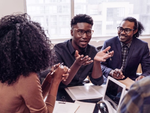 Diverse team collaborating around a table in a bright office setting, with one person standing and speaking enthusiastically to others.